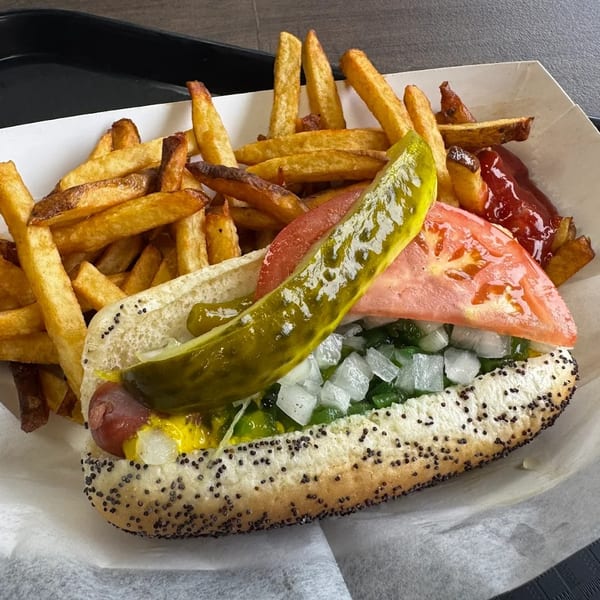 A Chicago-style hot dog with fries, ketchup, and a pickle spear in a paper tray.