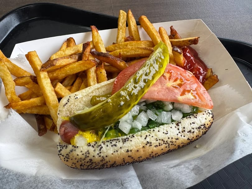 A Chicago-style hot dog with fries, ketchup, and a pickle spear in a paper tray.