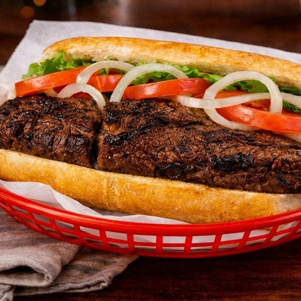 Grilled steak sandwich with lettuce, tomatoes, and onions in a red basket on a wooden table.
