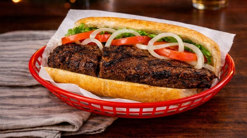 Grilled steak sandwich with lettuce, tomatoes, and onions in a red basket on a wooden table.